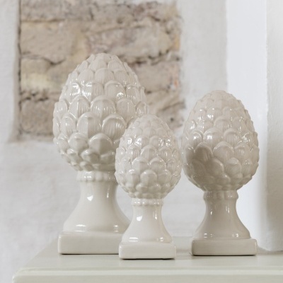 Três objetos decorativos em cerâmica branca em forma de pinhas sobre superfície e fundo branco com pedra