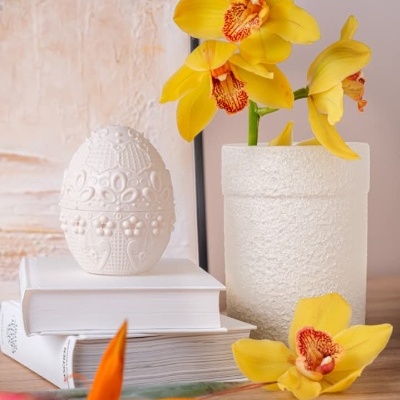 Vaso branco com flores amarelas, ovo decorativo e livros brancos sobre mesa de madeira