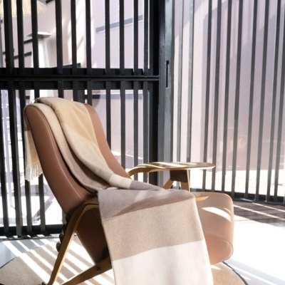 Cadeira de pele castanha com manta de lã bege, castanho claro e branco, em ambiente iluminado com janelas de ripas pretas