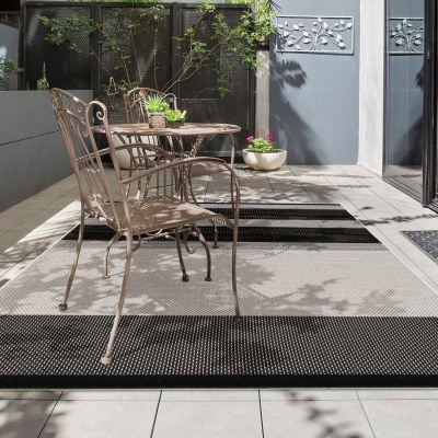 Mobiliário de exterior em metal castanho envelhecido com mesa e cadeiras, sobre tapetes cinza com padrão de pontos, numa varando com chão de azulejos claros e paredes cinza.