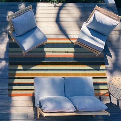 Conjunto de mobiliário de exterior com cadeiras e sofá de madeira clara e almofadas cinzentas sobre tapete listrado e pavimento em madeira