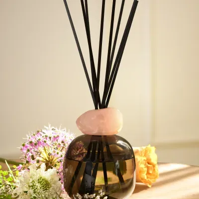 Frasco de difusor de ambiente com pedra rosa e varetas pretas, rodeado de flores coloridas