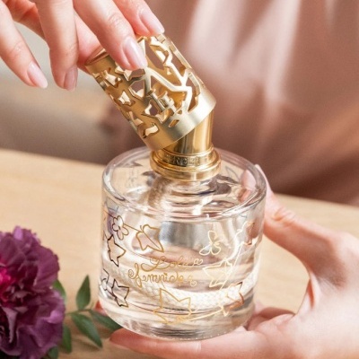 Frasco de perfume decorado com tampa dourada e desenhos dourados, mãos femininas seguram-no sobre mesa com flor roxa