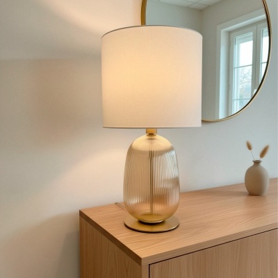 Candeeiro de mesa dourado com cúpula branca em móvel de madeira clara junto a espelho redondo.