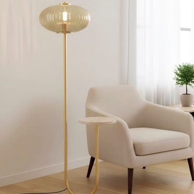 Lâmpada de pé dourada com abajur de vidro cinzento e mesa lateral pequena ao lado de cadeira bege em sala clara