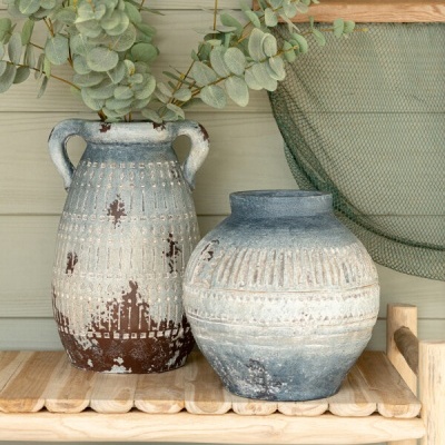 Dois vasos decorativos em cerâmica envelhecida com folhas verdes artificiais em prateleira de madeira clara