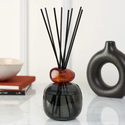 Difusor de aroma em vidro preto com aro âmbar e varetas pretas sobre superfície branca
