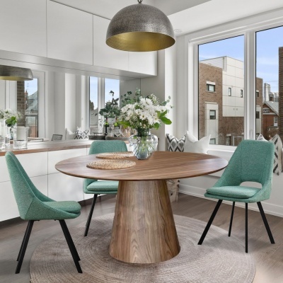 Mesa de jantar redonda de madeira com três cadeiras verde-água numa sala moderna com candeeiro pendente e flores no centro da mesa.
