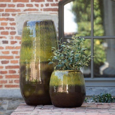 Vasos cerâmicos verdes e castanhos com planta sobre muro de tijolos junto a parede de tijolos e janela.