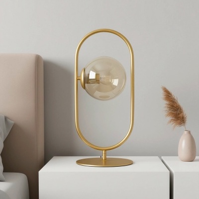 Candeeiro de mesa dourado com vidro esfumado numa mesa branca ao lado de uma cama e vaso com planta seca