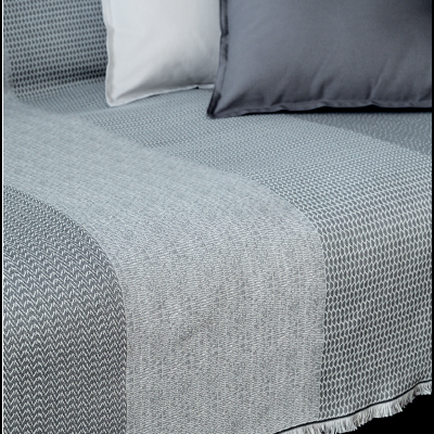 Colcha cinzenta com padrões e franjas, duas almofadas sobre sofá ou cama