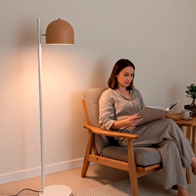 Lâmpada de pé branca com abajur terracota ao lado de cadeira de madeira clara com mulher a ler