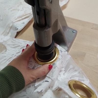 Mão com unhas vermelhas segurando tecido branco com padrão floral enquanto usa equipamento metálico para aplicar ilhós dourados