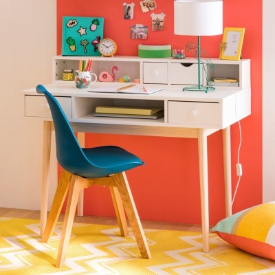 Mesa de estudo branca com cadeira azul e lâmpada, em ambiente com parede laranja e decoração colorida.