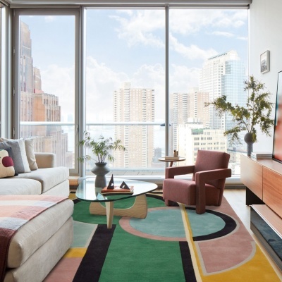 Sala de estar com sofá creme, poltrona rosa, mesa de centro de vidro, tapete geométrico e vista para cidade