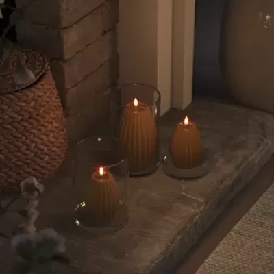 Três velas castanhas acesas em copos e base de vidro sobre pedra