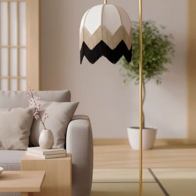 Luminária de pé dourada com cúpula branca e franjas bege e preto numa sala de estar com sofá cinza e planta