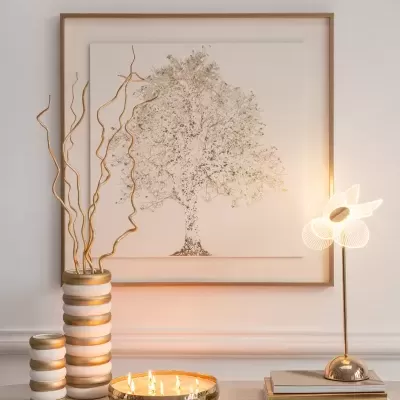Mesa com decoração branca e dourada, castiçal com velas acesas, candeeiro flor dourado e quadro com desenho de árvore dourada.