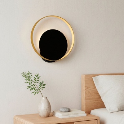 Luminária circular dourada e preta numa parede branca acima de mesa de cabeceira em madeira clara com vaso, livros e pedra, cabeceira de cama à direita.