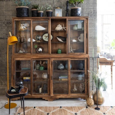 Aparador de madeira escura com portas de vidro e objetos decorativos, plantas em vasos e candeeiro de pé amarelo