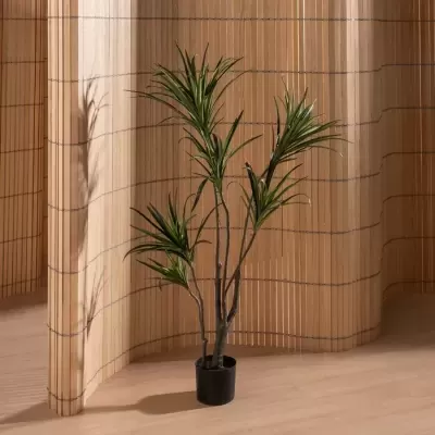 Planta artificial dracena em vaso preto, sobre chão de madeira clara, com ecrã de bambu ao fundo