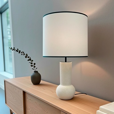 Candeeiro de mesa com base cerâmica branca e abat-jour branco sobre móvel de madeira clara com vaso preto e livros