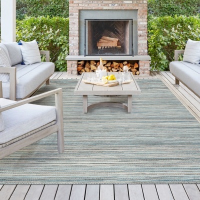 Área de estar exterior com tapete cinza, sofás de madeira com almofadas brancas, mesa de centro e lareira de tijolos
