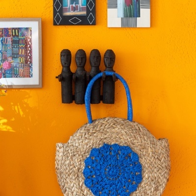 Bolsa redonda de palha com crochê azul pendurada em cabide de figuras esculpidas em parede laranja cercada por quadros artísticos.