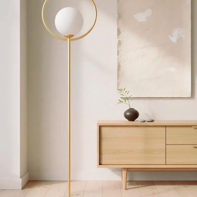 Candeeiro de pé dourado com bola branca e anel, junto a móvel de madeira clara e vaso com planta