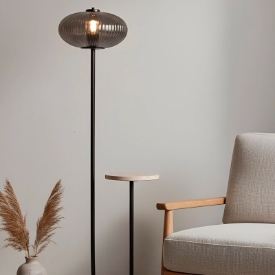 Candeeiro de pé com cúpula em vidro cinzento estriado e mesa lateral integrada ao lado de cadeira estofada cinza