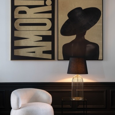 Duas obras de arte emolduradas numa parede, poltrona branca texturizada e luminária com base dourada e abajur preto