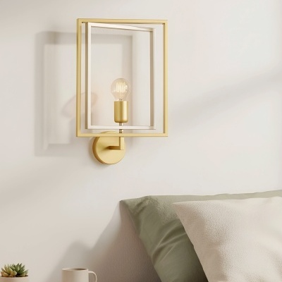 Candeeiro de parede dourado com estrutura quadrada acima de cama e mesa de cabeceira com caneca e livro.