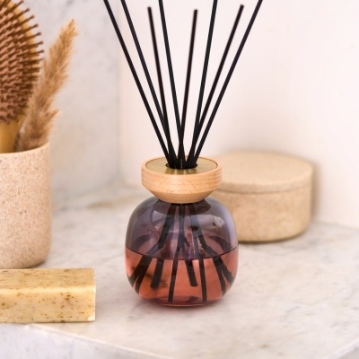 Frasco difusor de aroma em vidro fumado com tampa de madeira e paus pretos, sobre mármore clara, ao lado de sabonete e escova.
