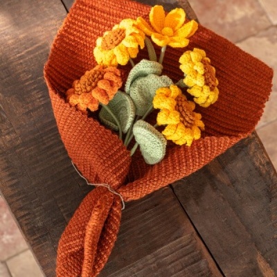 Bouquet de flores de croché amarelas e alaranjadas com folhas verdes em tecido laranja numa mesa de madeira.