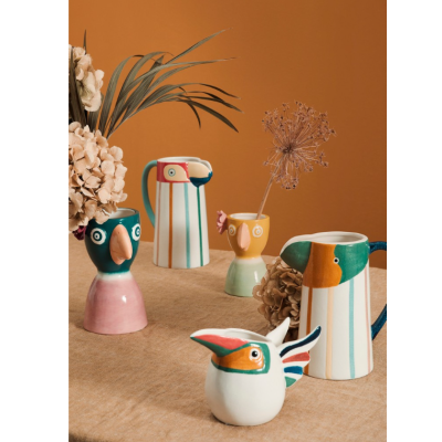 Cinco jarros cerâmicos decorativos em formas de pássaros, cores vibrantes, sobre mesa com toalha bege e fundo laranja.