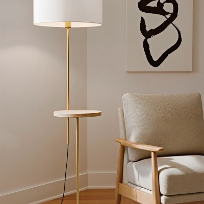 Luminária de pé dourada com abajur branco e prateleira de madeira ao lado de poltrona bege em ambiente neutro