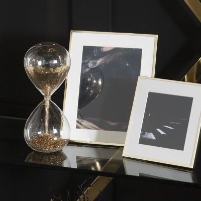 Relógio de ampulheta com areia dourada e duas molduras douradas com imagens abstratas sobre mesa de vidro