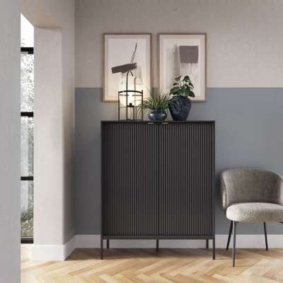 Móvel preto canelado com plantas e candeeiro, em parede cinza e bege com arte, ao lado de cadeira cinza.