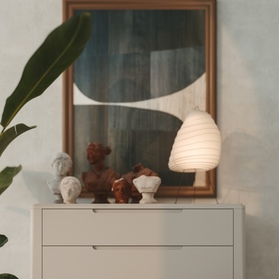 Cómoda branca com esculturas de bustos, lâmpada, quadro abstrato, planta e banco de madeira.