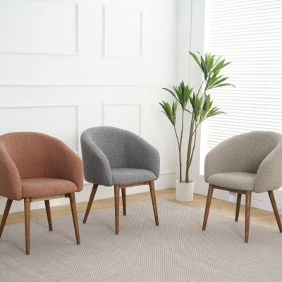Três cadeiras estofadas em tecido com pernas de madeira, sobre tapete cinza, planta verde em vaso e parede branca.