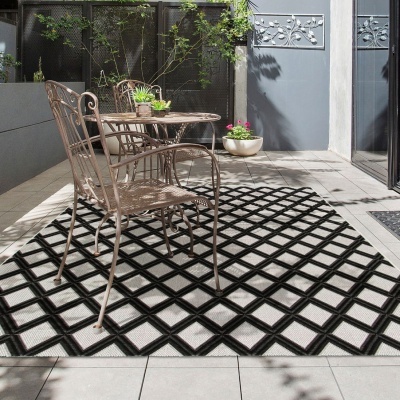 Conjunto de cadeiras metálicas e mesa redonda sobre tapete geométrico preto e branco num terraço externo com vasos e plantas.