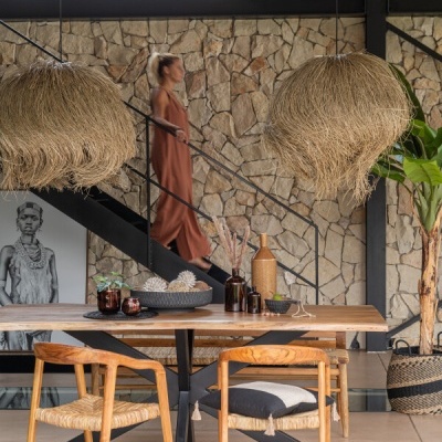 Mesa de madeira com cadeiras e luminárias de fibras naturais numa sala moderna com parede de pedra.