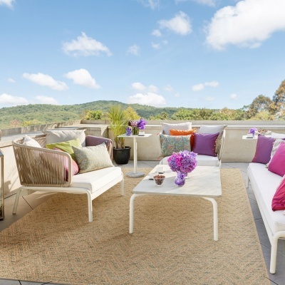 Móveis de exterior brancos com almofadas coloridas e mesa com flores lilás no tapete bege