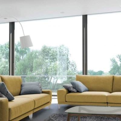 Sala moderna com dois sofás amarelos, almofadas cinza, mesa de centro de vidro, lareira suspensa e prateleiras pretas junto a grandes janelas