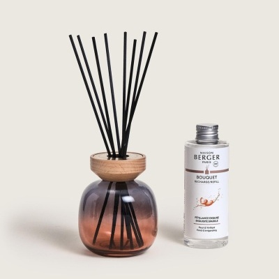 Aromatizador de ambiente em vidro rosé com varetas pretas e tampa de madeira, ao lado frasco de recarga transparente com rótulo branco.