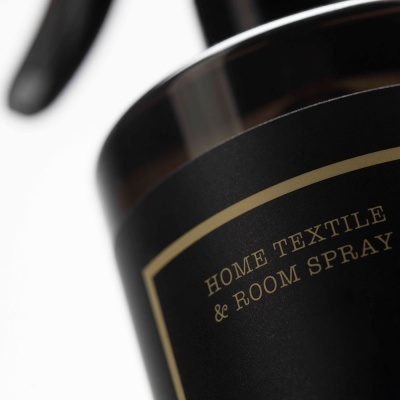 Frasco preto com rótulo dourado e texto 'HOME TEXTILE & ROOM SPRAY' em fundo branco