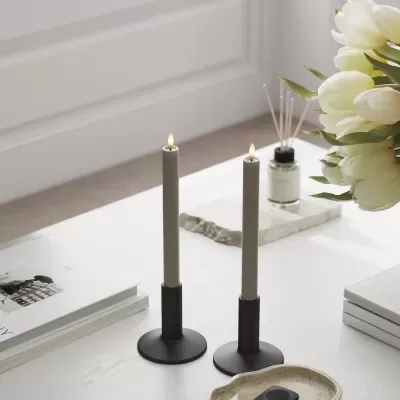 Duas velas cinzentas acesas em castiçais pretos numa mesa branca com flores, livros e aromatizador