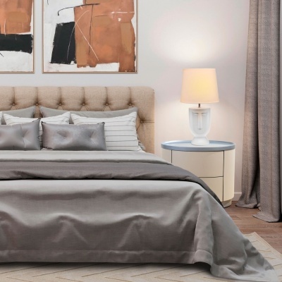 Cama estofada bege com almofadas cinzentas e brancas, mesa de cabeceira com candeeiro de rosto ao lado, quadro abstrato na parede.