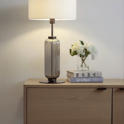 Lâmpada de mesa cinza com riscas verticais com cúpula branca sobre móvel de madeira com vaso de flores brancas e livros.