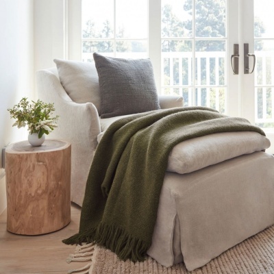 Poltrona bege com almofada cinza e manta verde ao lado de mesa rústica com vaso de flores